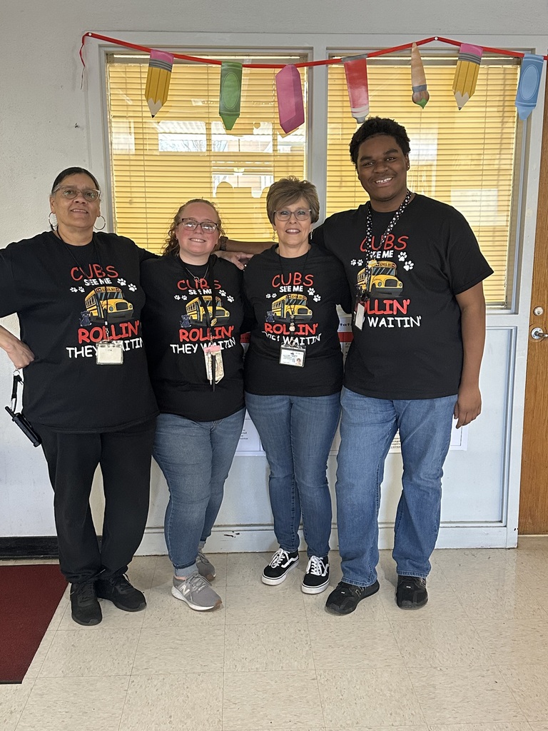A group photo of four adults — three women and one man — standing shoulder-to-shoulder and smiling. They wear matching black school-themed T‑shirts showing a yellow school bus with paw prints and a kitten peeking from a window. Above them, a string of colorful paper cutouts shaped like pencils and crayons hangs in front of a large window with horizontal blinds. The room has light-colored tile flooring with a red mat on the left and a light-colored door on the right. Visible text on the shirts/readable signage includes: "CUBS", "SEE ME", "ROLLIN'", and "THEY WAITIN'".