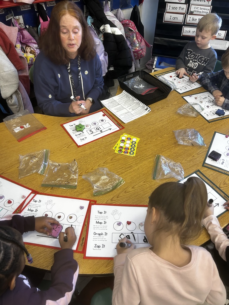 A teacher sits at a small group table with several young students during a literacy activity. The students use dry-erase markers and small manipulatives on laminated worksheets labeled with tasks such as “Tap It,” “Map It,” “Graph It,” and “Zap It.” Clear plastic bags and classroom materials are spread across the table while the teacher guides the lesson.