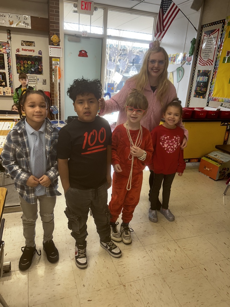 A blonde teacher wearing glasses and a pink sweater stands behind four young students in a brightly lit classroom. The children are dressed festively, with one boy wearing a red stylized "100" t-shirt, another child wearing red-framed glasses and multiple pearl necklaces, and a girl in a red shirt with a heart design. In the background, classroom elements are visible including a February calendar with dates 1-26, an alphabet chart showing "Ww Xx Yy Zz," an American flag, an EXIT sign above a door marked "47," a "Pledge of Allegiance" poster, a "MINDSET IS EVERYTHING" poster, and a "COOL KIDS" sign. Another student is partially visible seated at a desk in the far left background.