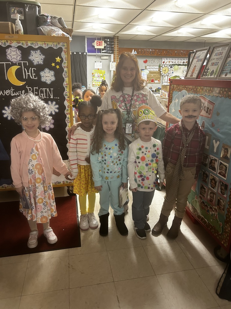 Seven people—six children and an adult teacher—pose in a classroom dressed in creative costumes for a "100 Days of School" celebration. Children are dressed as elderly characters, a sparkly astronaut, and other creative outfits, many holding books. The teacher wears a white shirt with a large "100" graphic and colorful checkered hearts. Behind them, a blackboard reads "TO THE adventure BEGIN..." with student names listed. Classroom posters display the alphabet, sight words, and motivational phrases like "Our little SEEDS grow mighty TREES." An EXIT sign is visible, and a red rug is on the tiled floor.