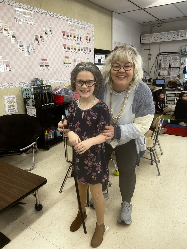 A smiling adult woman with blonde hair, glasses, and pearl necklaces stands with her arm around a young girl in a classroom. The girl is dressed in costume as an elderly person, wearing a gray wig, chain-link glasses, pearl necklace, dark floral dress, light brown leggings, and brown ankle boots while holding a black cane. The woman wears a light blue and white striped cardigan, dark pants, and light blue sneakers. Behind them, the classroom features a light pink patterned bulletin board displaying "VOWEL BABIES" and "CONSONANTS" with phonics cards, a poster reading "YOU CAN DO HARD THINGS," a dark circular chair, a small wooden desk, black storage bins labeled "QUIET BOOK," and other children visible in the background.