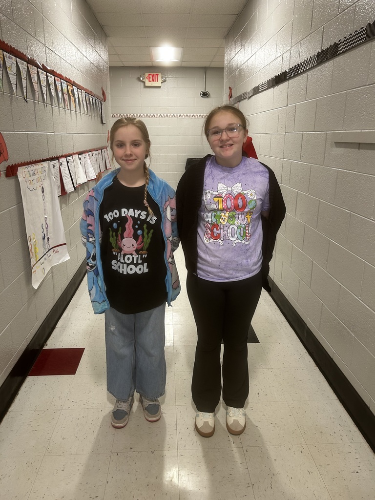 Two young girls stand side-by-side in a school hallway smiling at the camera. The girl on the left wears a black t-shirt with an axolotl design and "100 DAYS IS ALOTL SCHOOL" text, paired with a light blue and pink fleece jacket and colorful sneakers. The girl on the right wears glasses and a purple tie-dye t-shirt with "100 DAYS OF SCHOOL" text, paired with a black cardigan. Behind them, the hallway features light-colored tiled floors, white block-patterned walls decorated with student artwork with red accents, and an EXIT sign visible above.