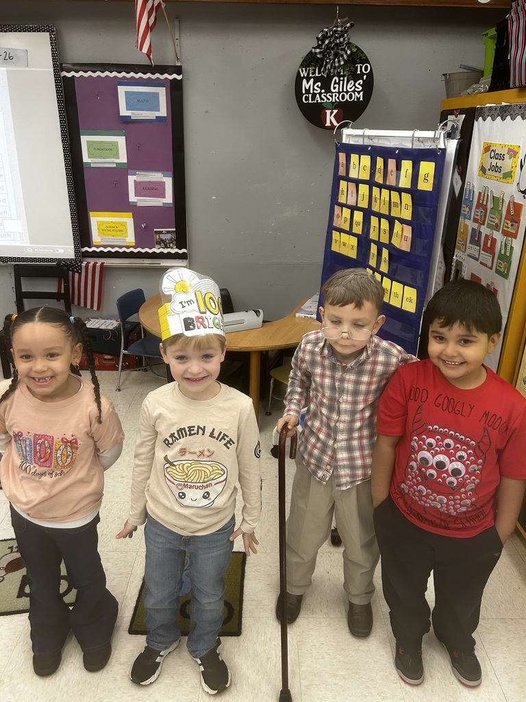 Four young kindergarten students celebrate the 100th day of school in a classroom. From left to right: a girl wearing a shirt reading "100 days of school" with gift box graphics; a boy wearing a hat reading "I'M [sun graphic] 100 BRIGHT"; a boy wearing a "RAMEN LIFE" shirt dressed as an elderly person with a plaid shirt, glasses, and walking stick; and a boy wearing a shirt reading "GOOD GOOGLY MOOD 100 DAYS OF SCHOOL" with googly eye graphics. Behind them, the classroom displays an American flag, a purple bulletin board labeled with "MATH FOUNDATIONS," "READING," and "SCIENCE/SOCIAL STUDIES," a circular welcome sign reading "WELCOME TO Ms. Giles' CLASSROOM K," a blue alphabet chart, and a "Class Jobs" chart.