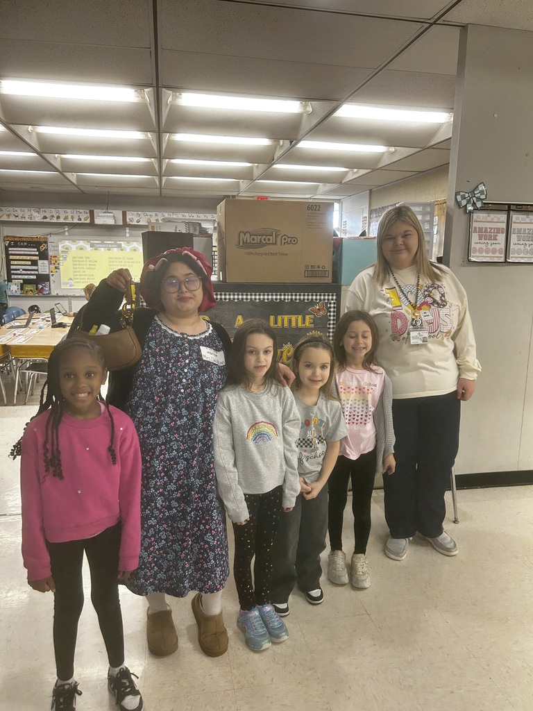 Five young elementary students and two adults pose together in a classroom. One adult is dressed in a floral dress with a red bonnet and glasses, holding a handbag, while another staff member stands beside the group wearing a white graphic shirt and lanyard. The children smile at the camera, wearing casual school clothes in bright colors. Classroom tables, chairs, posters, and storage bins are visible in the background.