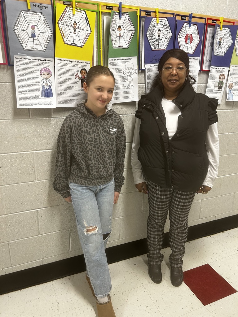 An elementary school student and a female staff member stand side by side in a school hallway. The student wears a gray leopard-print hoodie, light-wash ripped jeans, and brown boots, smiling slightly at the camera. The staff member wears a white long-sleeve shirt, black puffer vest, plaid pants, and black shoes. Behind them, colorful student projects about notable historical figures are clipped to a bulletin board on a cinderblock wall.