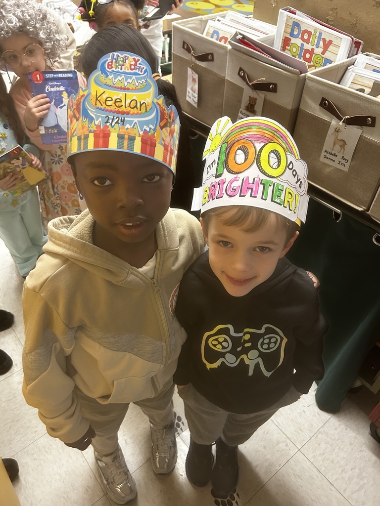 Two elementary students stand close together in a classroom, smiling at the camera and wearing paper celebration hats. One child wears a colorful “Happy Birthday” crown with the name “Keelan” written on it. The other child wears a hand-colored paper hat that reads “100 Days Brighter!” with a rainbow design. Classroom storage bins and folders are visible behind them.