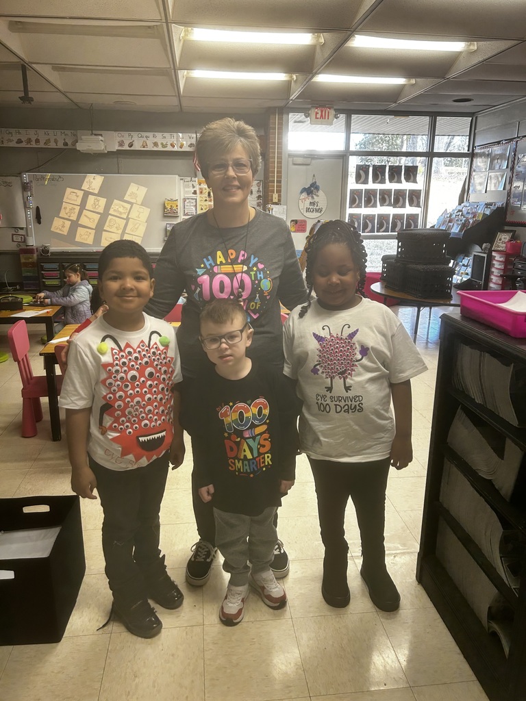 A teacher and three young children pose together in a classroom for a "100 Days of School" celebration. The teacher wears a grey shirt reading "HAPPY 100TH" with educational symbols. The children wear festive shirts: one with googly eyes and a monster mouth, one with "100 DAYS SMARTER" in colorful letters and pencil designs, and one with a purple monster graphic reading "I'VE SURVIVED 100 Days." Behind them, a whiteboard displays alphabet letters with picture cards, an EXIT sign is visible, and a nameplate reads "MRS. DECENOR." The brightly lit classroom has tiled ceilings, a pink table, storage bins, and educational displays.