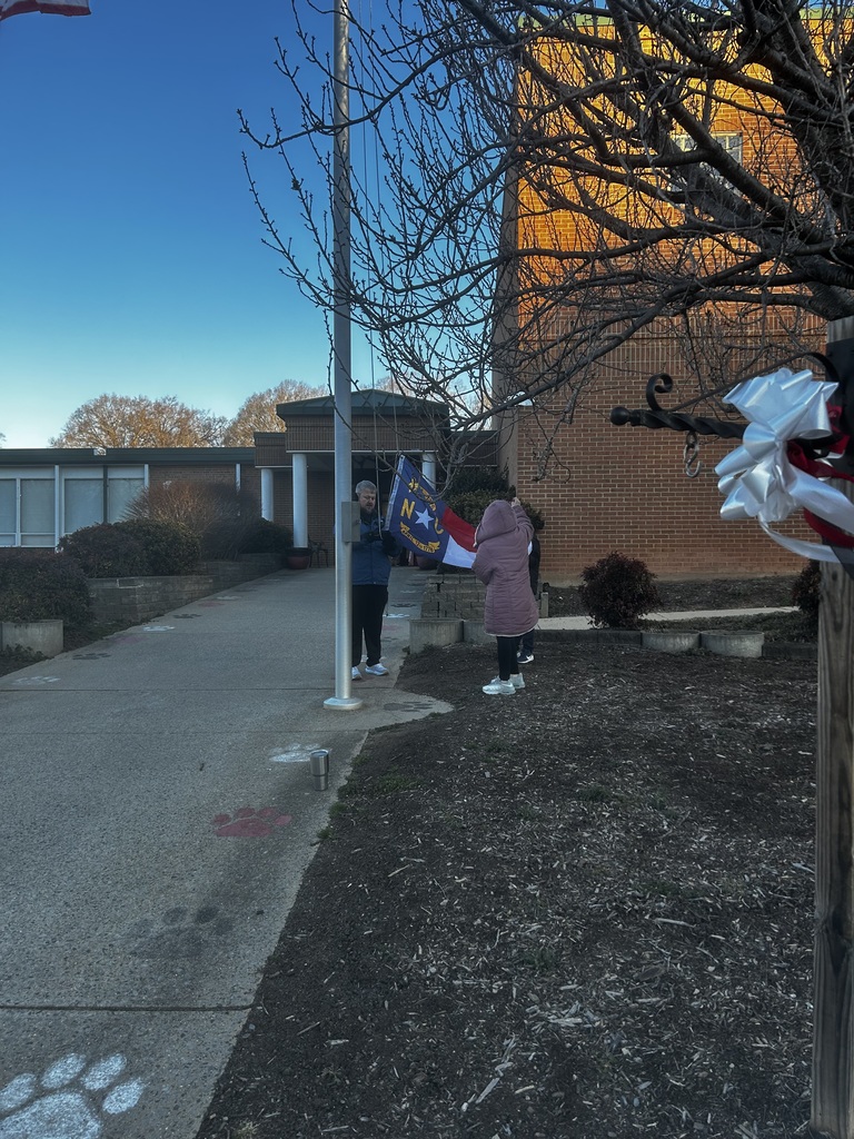 An adult in a blue jacket and a child in a brown hooded coat stand at the base of a flagpole in front of a brick school building, raising a North Carolina state flag. The flag displays the dates "MAY 20TH 1775" and "APRIL 12TH 1776" along with "N * C." Two other children are visible in the scene: one child with a backpack walks away on the left, while another child in a brown hooded jacket with a camouflage backpack runs on the right. Red paw prints are painted on the concrete pathway. The warm lighting suggests early morning or late afternoon, casting a golden glow on the brick building's wall.