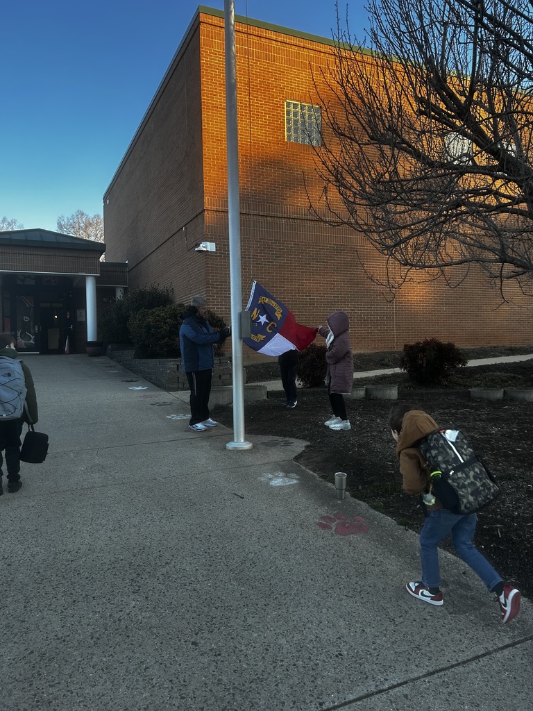 Two adults prepare to raise or lower a North Carolina state flag at an outdoor flagpole in front of a brick school building. A man in a blue jacket stands near the flagpole base while a woman in a purple/pink long coat holds the flag with him, with a third person partially visible behind them. The North Carolina flag displays a vertical blue stripe with a gold star, the letters "N C," and two banners showing the dates "MAY 20TH 1775." In the background, the brick building features large windows, white pillars supporting a covered entrance, and is illuminated by warm sunlight. Bare tree branches are visible on the right side. A concrete pathway with red and white painted paw prints leads toward the building, with a wooden post decorated with white and red/black bows visible on the right edge. The sky is clear and blue.