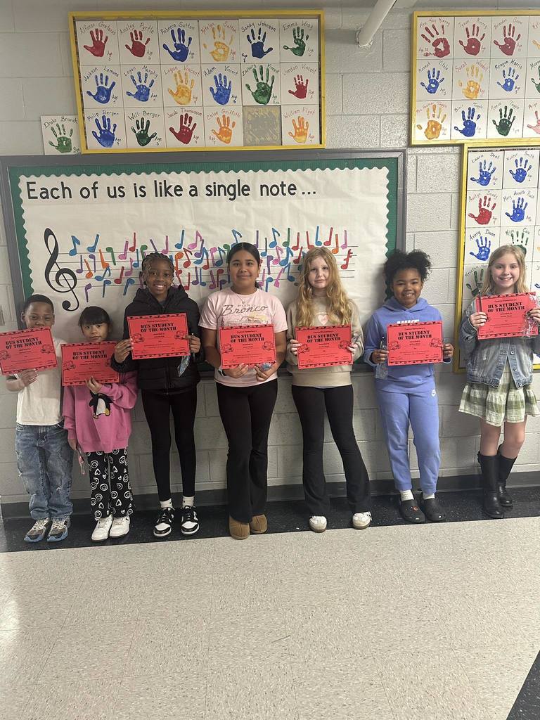 Seven elementary school students stand in a school hallway holding red “Bus Student of the Month” certificates. They are lined up in front of a bulletin board that reads, “Each of us is like a single note …” decorated with colorful musical notes. Above them, framed artwork displays painted handprints with students’ names. The students smile proudly while posing for the photo.