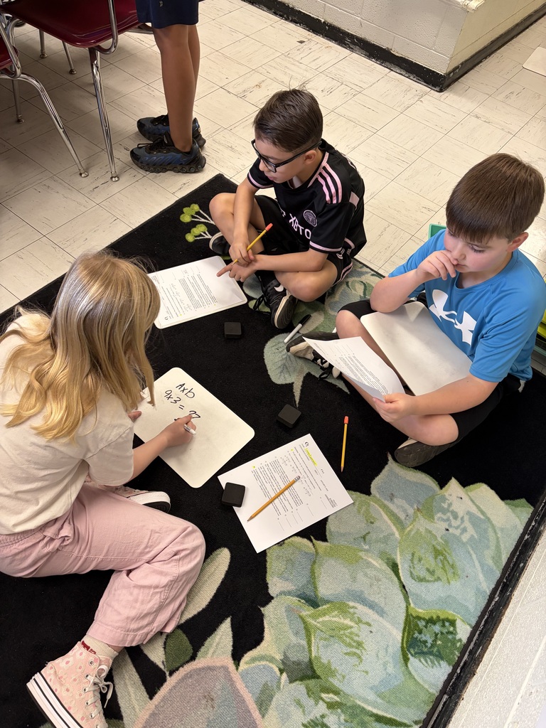 Three children sit on a black rug patterned with green and light green succulent designs, engaged in a learning activity in a classroom. A blonde girl wearing pink pants and white high-top sneakers with pink polka dots writes on a small white dry-erase board displaying "AxD 9x3 = 27 Comprehension." One boy wearing a black and pink striped soccer jersey with glasses holds a pencil while looking at a worksheet. Another boy wearing a blue t-shirt with an Under Armour logo also studies a worksheet. Pencils and small black erasers are scattered on the rug among their papers. In the upper left background, part of an adult's legs and a red classroom chair are visible along with a light-colored tiled floor.