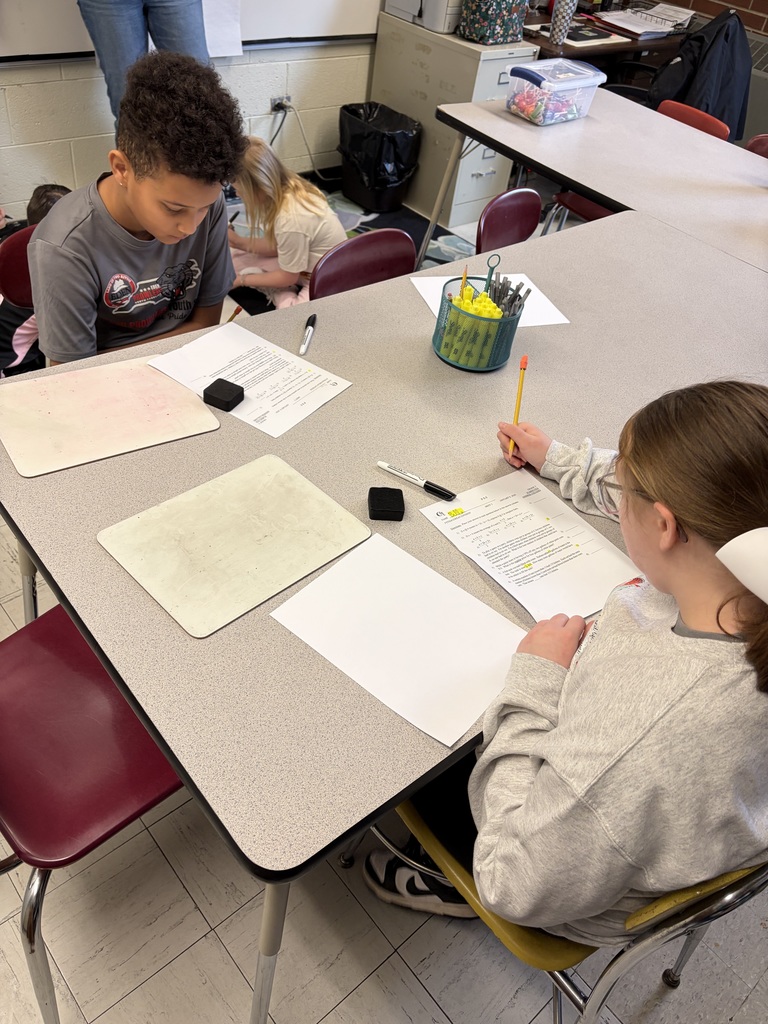 Two children work at a gray table in a classroom. On the left, a boy with dark curly hair wearing a "South Pride" graphic t-shirt looks down at his paper. On the right, a girl with light brown hair and glasses wearing a gray sweatshirt holds a pencil and writes on her paper labeled "Plate Boundaries Practice," containing twelve questions about plate tectonics, boundaries, and geological features. Between them on the table are two small whiteboards, two black markers, two black erasers, a blank white paper, and a teal mesh pencil holder filled with yellow pencils and writing tools. In the background, another child with blonde hair works on the floor. An adult's legs are partially visible in the upper left corner. Gray metal filing cabinets, additional tables, chairs, a black trash can, and colorful supply containers are visible throughout the classroom with a tiled floor.