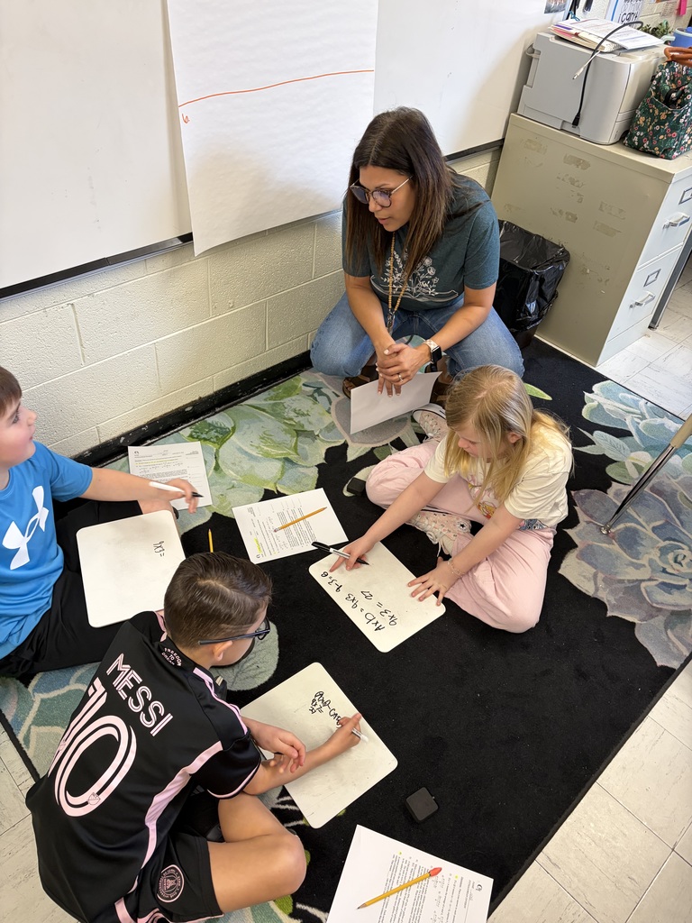 A teacher wearing glasses and a dark green shirt kneels on a floral-patterned rug with three students sitting around her on the floor. The students—two boys and one girl—are engaged in a learning activity using small whiteboards with markers. One whiteboard shows "4x3 = 27," another displays "AND OR x+y," and a partially visible whiteboard shows "4x3 =." The girl wears a light-colored shirt and pink pants. One boy wears a blue shirt with an Under Armour logo, and the other boy wears a black and pink Messi #10 jersey. Papers, pencils, and markers are scattered on the rug. In the background, a white brick wall displays a large paper chart, filing cabinets, and a trash can are visible.