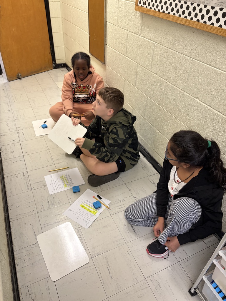 Three children sit on a light-colored tiled floor in a school hallway or classroom engaged in an activity. On the left, a boy wearing a peach-colored hooded sweatshirt with a crown design holds a pencil. In the middle, a boy wearing a camouflage hooded sweatshirt and black shorts holds a small whiteboard and marker while looking upward. On the right, a girl with glasses wearing a black jacket, white shirt with red detailing, and black and white checkered pants is tying her black and pink athletic shoe, with her dark hair pulled back in a ponytail with a green elastic band. Papers with printed text and some sections highlighted in yellow, small whiteboards, pencils, and blue erasers are scattered on the floor around them. A white shelving unit with clear bins is partially visible on the right, with a light beige brick wall and brown door visible in the background.