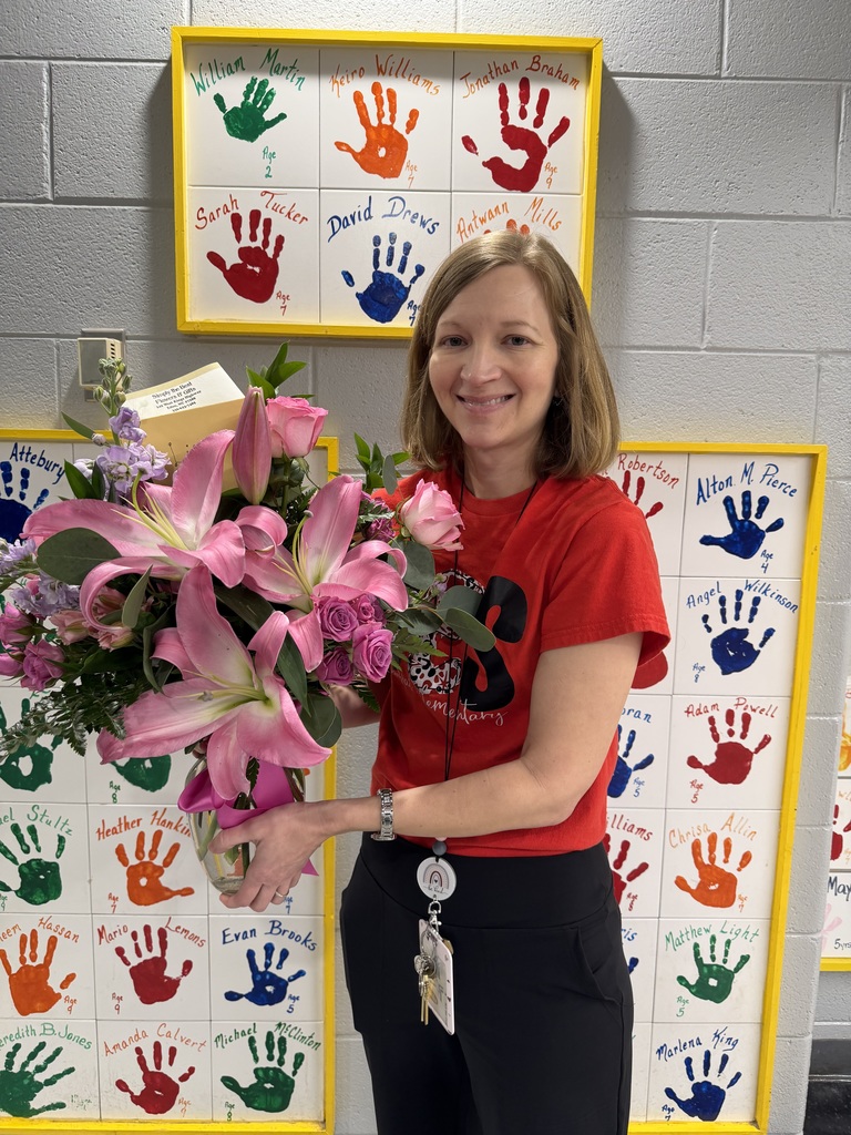 A smiling woman wearing a red T-shirt stands in a school hallway holding a large bouquet of pink lilies, roses, and greenery in a glass vase. Behind her are wall displays of colorful painted handprints on white tiles, each labeled with a student’s name and age, framed in bright yellow. She wears a lanyard with keys and an ID badge, and the hallway wall is made of light gray cinder blocks.