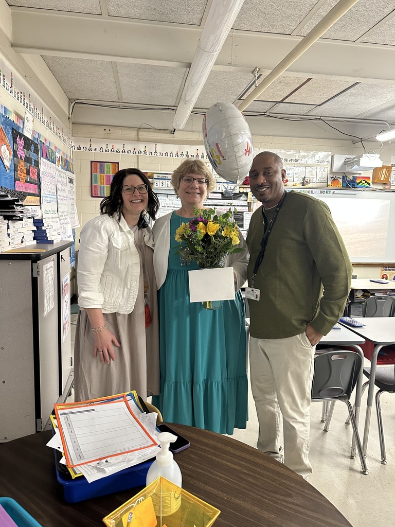 Three school staff members pose together in a classroom. The person in the center holds a bouquet of yellow flowers and a card, with a balloon floating above them. The two colleagues beside her smile while standing close. Desks, student charts, and classroom materials fill the background, creating a bright and welcoming classroom setting.