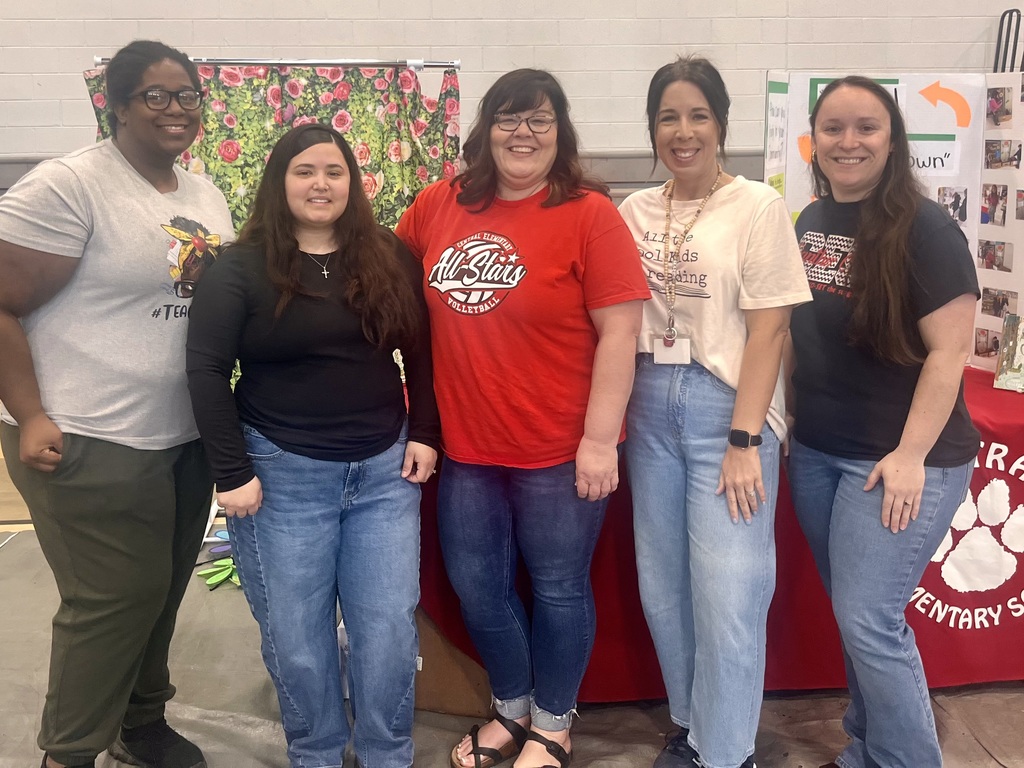 Five women stand side-by-side smiling at the camera at an indoor school event. From left to right, they wear: a light gray t-shirt with "#TEAC" (partially visible) and a person-with-books graphic, a red t-shirt reading "CENTRAL ELEMENTARY All-Stars VOLLEYBALL," a cream t-shirt reading "All the Cool Kids Reading" with an open book graphic, and a black t-shirt with partially visible text "EERS." The background features a light-colored wall, a floral curtain with red roses, and a white display board with pictures and text. A red table with a school-themed banner displaying a white paw print and "CENTRAL ELEMENTARY SCHOOL" text is visible in the foreground on the right, along with an orange arrow pointing right and the word "OWN" on the display board.