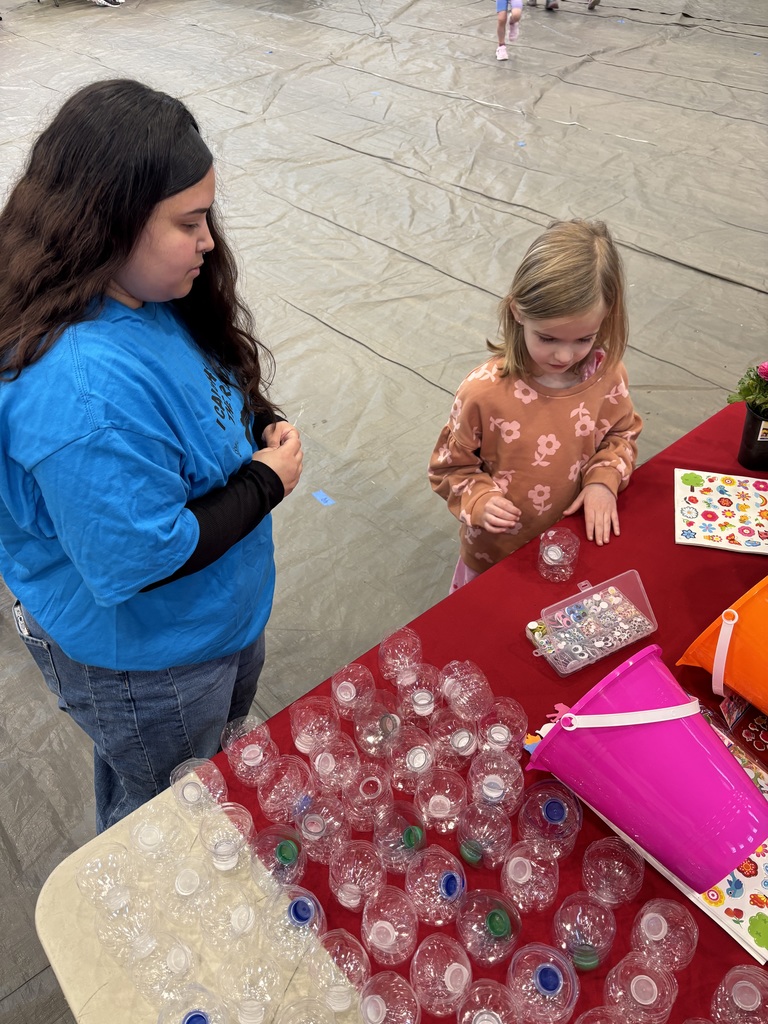 An adult woman with long dark hair wearing a blue t-shirt and jeans stands with a young girl with blonde hair wearing an orange long-sleeved shirt with flower patterns and pink pants at a red-draped table. On the table, numerous small clear plastic bottles with colored caps in blue, green, and white are arranged alongside a clear container filled with small shiny beads, charms, and rings. A pink bucket with a white handle, sheets of colorful stickers featuring hearts, stars, flowers, and nature-themed designs, and a small potted plant with pink flowers are also visible. The floor in the background features a large light-colored tarp or mat with faint lines, and other people, including children, are visible in the distance.