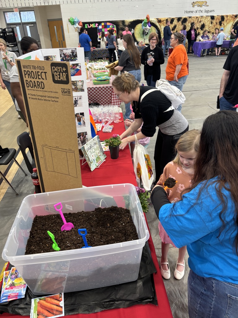 An indoor community event takes place in a large gymnasium hall with a "Home of the Jaguars" banner and jaguar mural visible on the back wall. Numerous attendees walk and interact with various booths displaying red and checkered tablecloths and balloons. In the foreground, a booth features a red-covered table with a large clear plastic bin filled with soil and small colorful plastic gardening tools in pink, green, and blue. Seed packets for Ferry-Morse Wildflowers and Carrots, a small potted plant, and a book titled "Harlem Grown" displayed on a stand are on the table. A woman in a black top arranges items while another woman helps a child scoop soil from the bin. A brown tri-fold project board with photos is displayed prominently on the table.