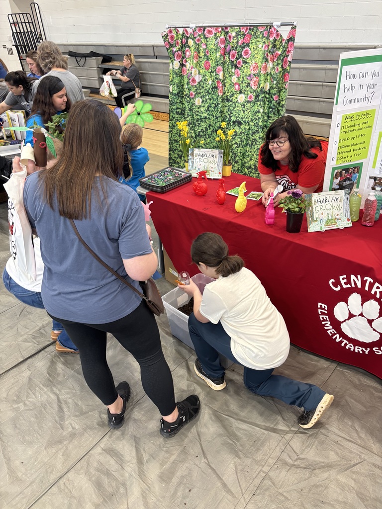 An indoor school community fair event is held in a gymnasium. A child in a white shirt and blue pants kneels on the floor, transferring soil from a clear bin into a small pot while a person in a blue shirt and black leggings observes. Behind them is a booth with a red tablecloth displaying "CENTRAL ELEMENTARY SCHOOL" with a paw print logo. A woman in a red shirt stands behind the table smiling. The booth features a green leafy and pink rose floral backdrop, with items including spray bottles in red, yellow, and pink, potted daffodils and purple plants, and multiple copies of the book "Harlem Grown." A display board on the right lists "Ways to help in your Community," including donating used toys, books and clothes, picking up litter, reusing materials, spreading kindness, and starting a free library.