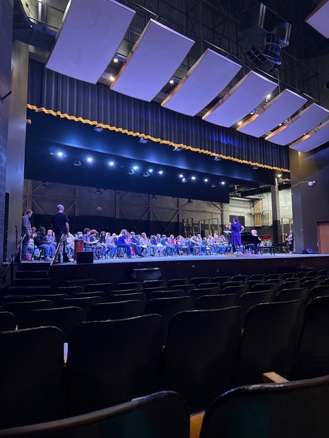 A wide shot of an auditorium or theater shows numerous dark, unoccupied seats in the foreground. On the illuminated stage with blue and purple lighting, a large group of people are seated in rows, arranged as an orchestra or choir during rehearsal. Many individuals hold instruments or music. A person in a purple dress stands prominently on the stage, facing the seated group and conducting or directing them. Another person is seated at a piano on the right side of the stage. The stage features visible stage lights across its front edge, and multiple large rectangular acoustic panels suspended from the ceiling above are bathed in purple hue from the stage lighting. A decorative golden wavy element runs along the top of the stage opening, and the ceiling structure above features exposed industrial elements.