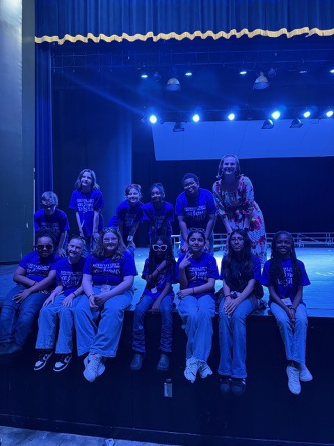 Twelve children and young teenagers and one adult woman pose together on a stage for a photo. Several children are seated on the edge of the stage in the foreground while others stand or crouch behind them. The adult woman, wearing a floral dress, stands on the right side of the upper group. The background features a dark blue stage curtain with visible stage lights above, creating a blue-tinted lighting effect.