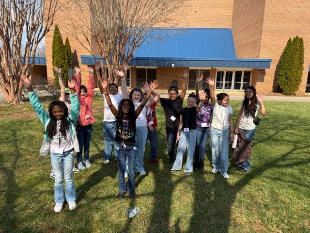 Twelve elementary or middle school-aged children stand on a sunny, grassy lawn in front of a brick building with a blue roof and several bare trees. Most of the children are smiling with their arms raised overhead, waving or gesturing enthusiastically. They are dressed in casual clothing including jeans and t-shirts. Long shadows of the children are cast on the grass in front of them, indicating bright sunlight.