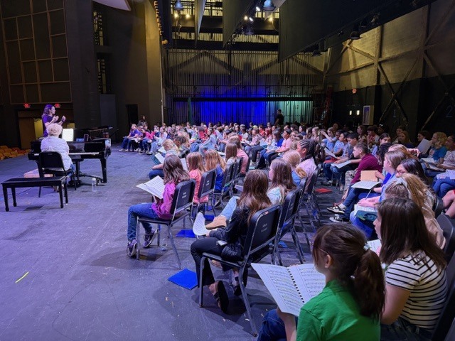 A large group of children, likely students, are seated in rows in an auditorium or rehearsal space on a stage. Most are holding sheet music or notebooks and facing toward the front. Two adults lead the group: one seated at a grand piano facing away from the camera and another standing with hands raised as if conducting or instructing. The space features a high ceiling with exposed structures and a dark curtain with blue lighting in the background, suggesting a music lesson, choir practice, or rehearsal session.