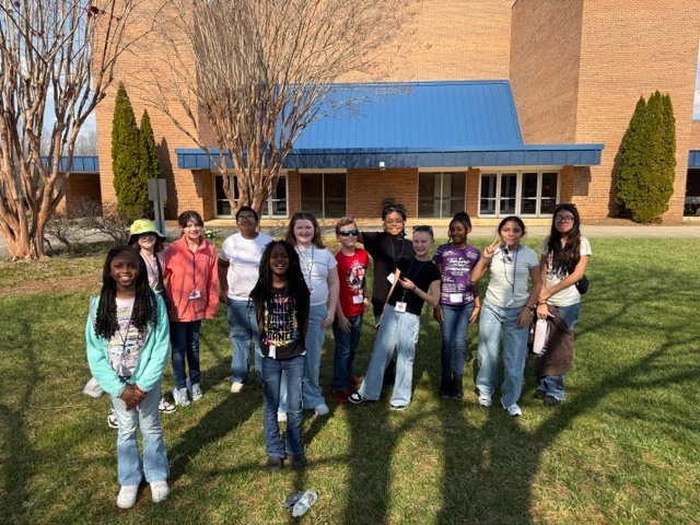  Approximately 12 diverse children and young teenagers stand outdoors on a sunny day on a grassy area in front of a brick building with a prominent blue sloped roof section. Several bare-branched trees are visible in the foreground and background, suggesting late winter or early spring. Most of the children are smiling and looking toward the camera, dressed in casual attire including jeans, t-shirts, and hoodies. Some children wear lanyards around their necks. Long shadows are cast on the grass from the people and trees.