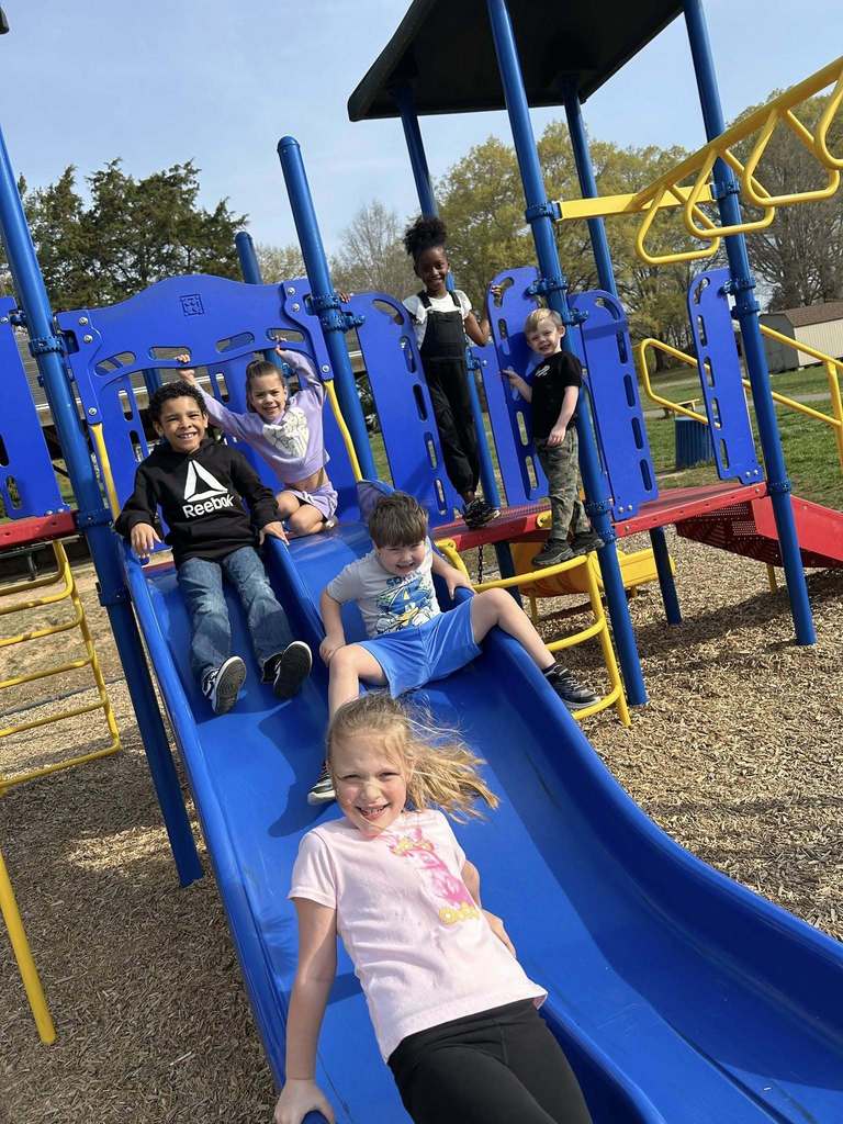 Six children actively play on a blue and yellow playground structure on a sunny day. In the foreground, a blonde-haired girl in a pink t-shirt and black leggings smiles while sliding down the blue slide. Behind her, a boy in a grey t-shirt and blue shorts sits on the slide. Further up, a boy wearing a black Reebok hoodie with a white delta symbol smiles while on the slide. Above him, a girl in a light purple outfit sits on the slide with her arms raised. On a red platform to the right stand two more children: a girl with dark curly hair wearing black overalls and a white t-shirt, and a boy in a black t-shirt and camouflage pants. The background shows green grass, several trees under a clear blue sky, and hints of other playground equipment and a building in the distance.