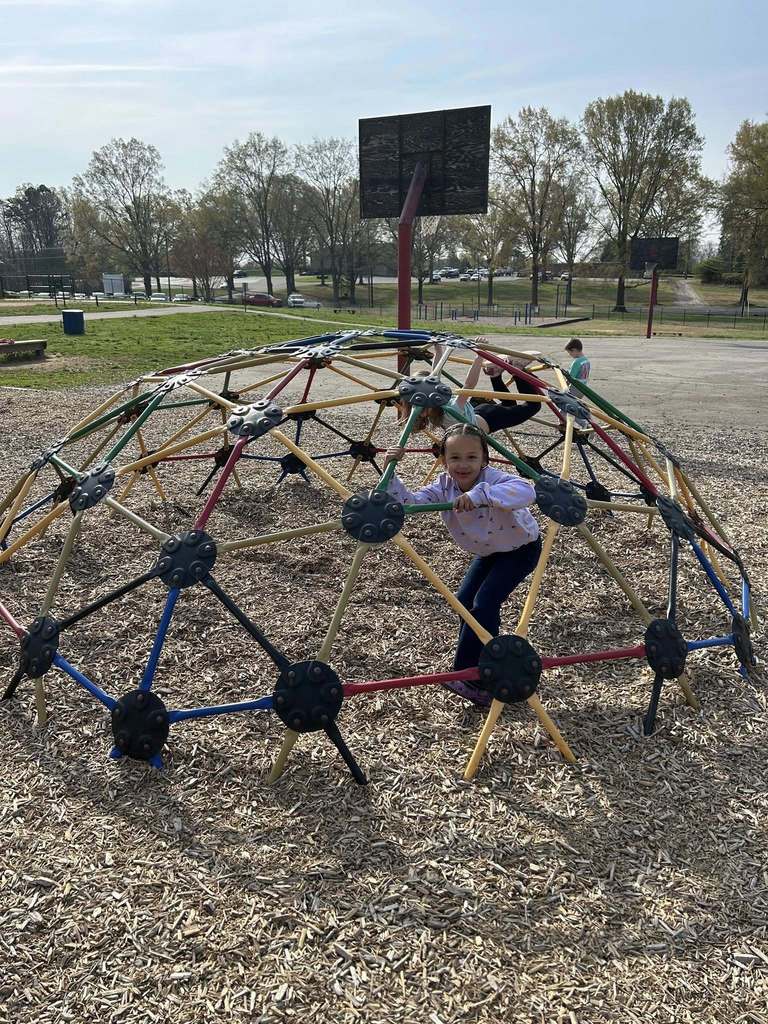 Several children play on a large, colorful geodesic dome climbing structure at a sunny outdoor playground. In the foreground, a young girl with braids wearing a light-colored long-sleeve patterned shirt and dark pants smiles directly at the camera while holding onto the dome's bars. Other children are visible further back and higher up on the dome: one in a dark shirt is partially visible, and another in a light-colored shirt is on the far right. The ground beneath the structure is covered with wood chips. In the background, a grassy field, trees with sparse leaves suggesting early spring or late fall, and two basketball hoops with backboards are visible. A parking lot with cars and buildings appear in the distance under a light blue sky.