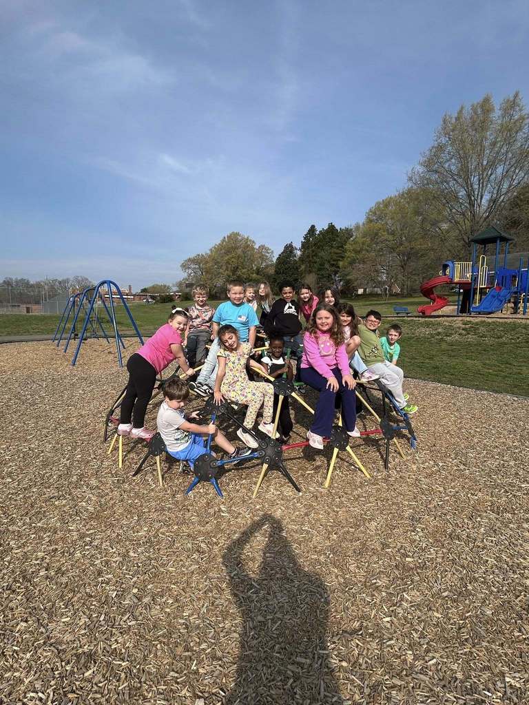 Approximately 15 elementary school-aged children gather on and around a black and yellow geometric climbing structure shaped like interconnected pyramids or tripods on a sunny playground day. Many children are smiling and looking toward the camera. A girl in a pink shirt and black leggings climbs on the left side while a boy in a grey shirt and blue shorts sits on a lower section. A girl in a pink hoodie sits prominently toward the front right. The playground features wood chip ground cover, with a blue swing set visible on the left in the background. Grassy areas, trees, and a larger colorful playground structure with red and blue slides are visible further back. The clear blue sky has faint wispy clouds. A shadow of the photographer holding a camera is cast on the wood chips in the foreground.