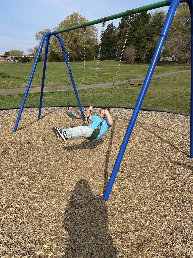 A child or young adolescent actively swings on a blue and green swing set in a park on a sunny day. They wear a bright blue t-shirt, light-wash ripped jeans, and black and white sneakers, with their body angled mid-swing and legs extended forward. Light brown wood chips cover the ground beneath the swing set. In the background, a large grassy area, numerous trees with varying degrees of foliage, a winding paved path, and a brown park bench are visible under a clear blue sky.