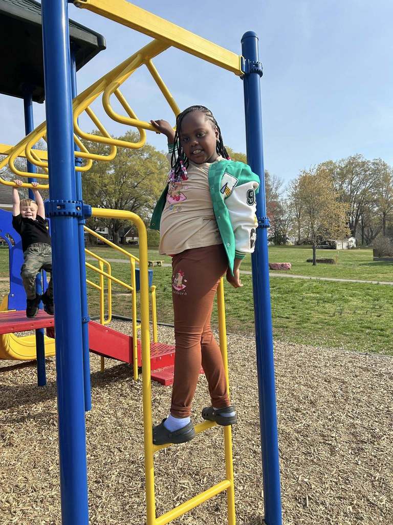 Two children play on a sunny playground. A young Black girl is in the foreground, climbing a yellow ladder on the playground structure. She wears a beige t-shirt, a green and white varsity-style jacket, brown leggings, dark grey clogs with light blue socks, and has beaded braids in her hair. She looks toward the camera with a slight smile. In the background, a young white boy hangs from the monkey bars. The playground features blue support poles, yellow bars and ladders, and red slides and platforms. Wood chips cover the ground, with a grassy area and trees visible under a clear blue sky.