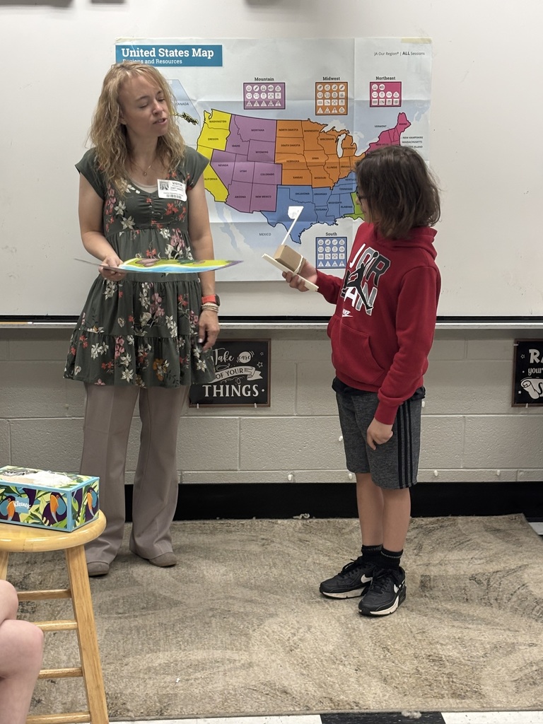 A woman wearing a floral dress and khaki pants stands with a child in a red hoodie and grey shorts in front of a large United States map displayed on a white classroom wall. The map, titled "United States Map: Regions and Resources" with the "JA Our Region | ALL Sessions" label, divides the country into four colored regions: Mountain (purple), Midwest (orange/yellow), Northeast (red), and South (blue/green). Each region displays state names and resource boxes containing six icons arranged in two rows of three with circle, square, and triangle patterns. The map shows borders with Canada to the north and Mexico to the south, and includes state labels throughout. The woman holds an open colorful book or poster. The child holds a small handmade object constructed from cardboard and sticks. A box of Kleenex tissues sits on a small wooden stool in the lower left foreground. The child wears a Nike hoodie with "JR AN" (part of Jordan) visible. A wall sign reads "Take care OF YOUR THINGS." The woman wears a "VISITOR" badge. The lower wall features a grey brick pattern, and the floor is carpeted with a light-colored rug.