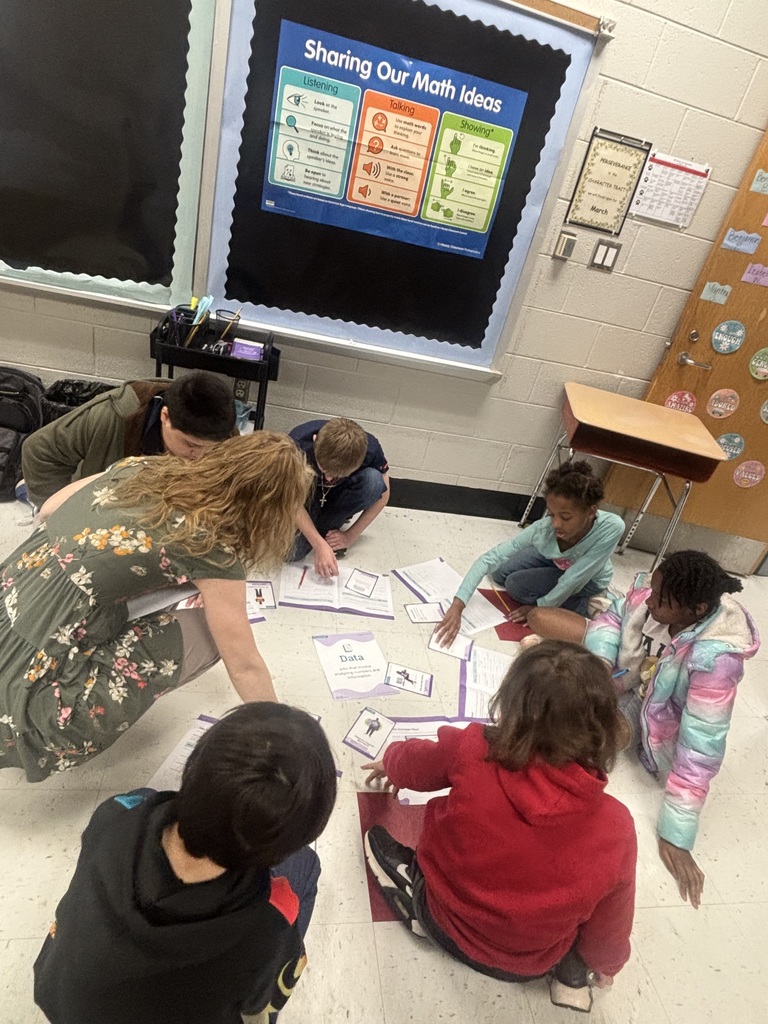 Six elementary school-aged children and one adult teacher sit on the floor in a classroom engaged in a math learning activity with several papers and workbooks spread around them. The adult leans forward, pointing at papers while children look at the materials and some hold pencils. A bulletin board behind them displays a "Sharing Our Math Ideas" poster with sections on Listening (look at the speaker, focus on what they're saying, think about their ideas, be open to new strategies), Talking (use math words, ask questions, use strong voice with class or quiet voice with partner), and Showing (I'm thinking, I have an idea, I agree, I disagree). A classroom door is decorated with numerous positive character trait words including "ENOUGH," "LOVED," "HELPED," "CALM," "READY," "LEARN," "LISTEN," "BRAVE," "cooperative," "kind," "friendly," "respectful," "gentle," "secure," "creative," "patient," "positive," "strong," "empathetic," "thoughtful," "fair," "peaceful," "curious," "honest," "generous," "leader," "sincere," "dependable," "organized," "diligent," "focused," and "responsible." A wall sign displays "PERSEVERANCE is the CHARACTER TRAIT for March." A paper on the floor shows text about "Data that involves analyzing information." A small desk is visible in the background.