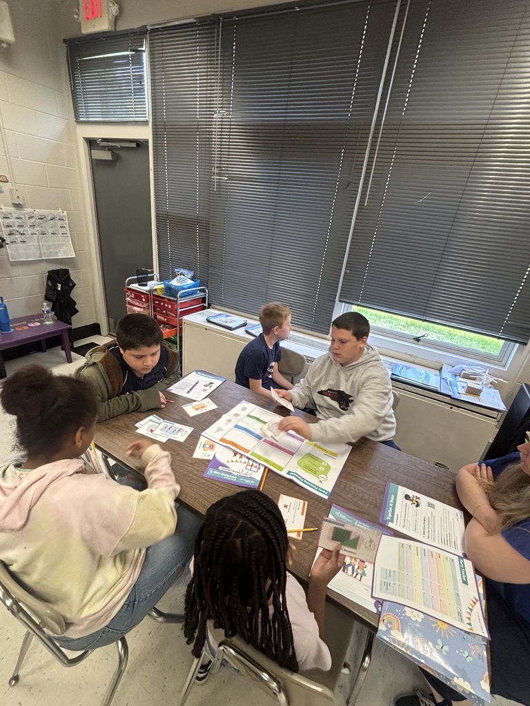 Five students and one adult engage in an educational activity around a rectangular table covered with colorful worksheets, activity guides, and small cards in a classroom. One student in a grey hoodie handles papers while another student with braids holds up a card. Documents on the table include "Party Planning Proposal," "Our Learning Log," "Together at Home," and "Party Details" with a "MATH" label visible on a small card. The classroom features a white brick wall on the left, a dark gray door with an "EXIT" sign above it, and large windows with dark gray horizontal blinds on the right. A red rolling storage cart and a small purple table are visible in the background.