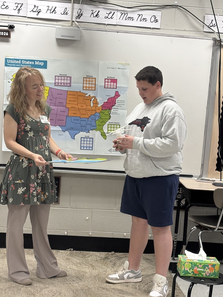 A woman wearing a floral dress stands with a boy in a grey hoodie with a black and red logo and blue shorts in front of a whiteboard displaying a large United States map titled "United States Map: Regions and Resources" with the "Junior Achievement" logo. The whiteboard also shows cursive letters "Ee Ff Gg Hh Ii Jj Kk Ll Mm Nn Oo" across the top and "2026" on the left side. The map divides the U.S. into regions—Mountain, Midwest, Northeast, South Atlantic, and South—each with four symbol icons (circle, square, X, triangle) representing resources. State labels are visible throughout the map, including Vermont, New Hampshire, Maine, Massachusetts, Rhode Island, Connecticut, New York, Pennsylvania, New Jersey, Delaware, Maryland, Washington D.C., Virginia, North Carolina, South Carolina, Georgia, Florida, and many others, with Canada and Mexico shown as borders. The woman holds a colorful board or poster. The boy holds a craft project made of straws and possibly cups. A box of Kleenex tissues with a floral design sits on a stool to the right. The text "JA Our Region | ALL Sessions" appears in the map's top right corner.