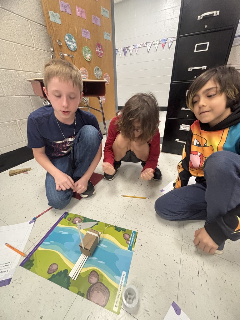 Three young boys sit on a tiled classroom floor engaged in a hands-on learning activity around a colorful "River Placemat" labeled "JA Our Region® | Session 3: Design." The boy on the left wears a blue t-shirt and jeans, kneeling while speaking or explaining something. The boy in the middle has curly hair and wears a red long-sleeved shirt, squatting low and intently focused on the craft. The boy on the right wears a colorful hoodie featuring a Charizard-like character and dark pants, sitting cross-legged with a thoughtful expression. Their craft project consists of a small brown cardboard box forming a boat's hull with several white cylindrical objects (straws) taped underneath as pontoons, a white paper sail attached to a vertical stick/straw mast, and a thin white string extending from the boat. Clear tape and an orange pencil sit beside the project, with a yellow pencil nearby. The background shows a white cinder block wall, a partial classroom desk with a red top, a wooden door or bulletin board decorated with student names on colored papers and circular affirmations reading "I AM ENOUGH," "I AM GENEROUS," "I AM KIND," "I AM BRAVE," "I AM COURAGEOUS," "I AM SPECIAL," and "I AM LOYAL."