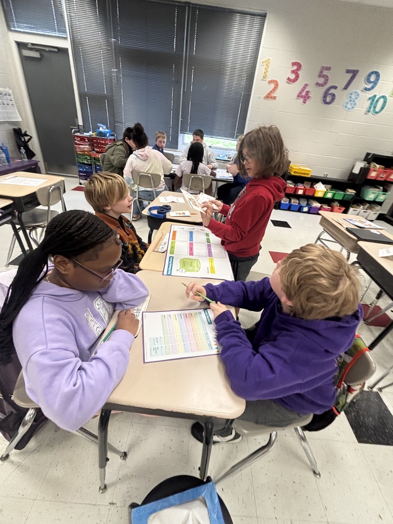 Several elementary or middle school-aged students engage in a learning activity in a brightly lit classroom with large windows featuring horizontal blinds. On one wall, colorful number cutouts are displayed in two rows: "1 3 5 7 9" and "2 4 6 8 10." In the foreground, three students are focused on their work: a student in a light purple hoodie and glasses sits at a desk on the left, writing on a worksheet with a pencil; a student in a dark purple hoodie sits at another desk, holding a pencil and looking up; and a student in a red hoodie stands between them, holding and examining a small booklet or set of cards. Desks display papers and activity boards with grid-like layouts containing multiple colored cells.
