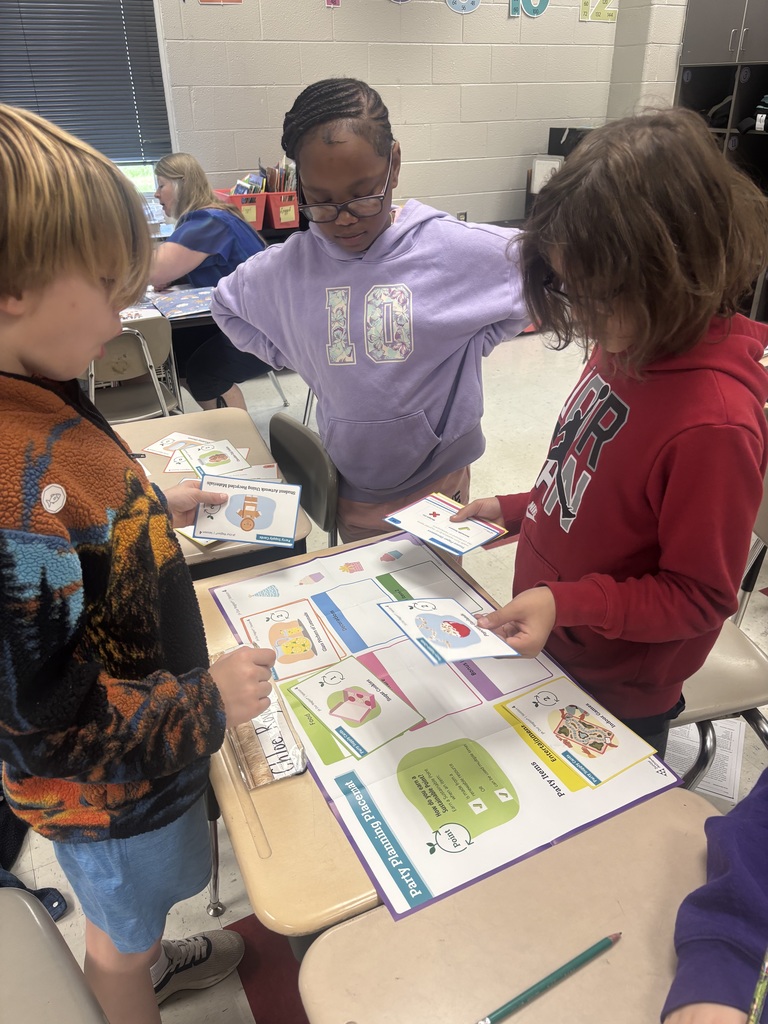 Three children engage in an educational activity around a table in a classroom, with an adult visible in the background at another table. The child on the left has blonde hair and wears a colorful patterned fleece jacket. In the middle, a girl with braided hair and glasses wears a purple hoodie. On the right, a child with brown hair and glasses wears a red hoodie. They work with a large "Party Planning Placemat" and small cards featuring party-related items. The placemat asks "How do you plan a birthday party?" and organizes party items into categories: Food (snacks, drinks, desserts), Entertainment (indoor games, outdoor games, crafts), and Goody Bags (balloons, treats, party favors). Visible cards include "Musical Chairs Using Recycled Materials" and "Blue Cupcakes with Red Cherries," along with illustrated cards depicting a yellow cake with pink frosting, a green drink, a pink present, balloons, and a person with a party hat and blue balloon.