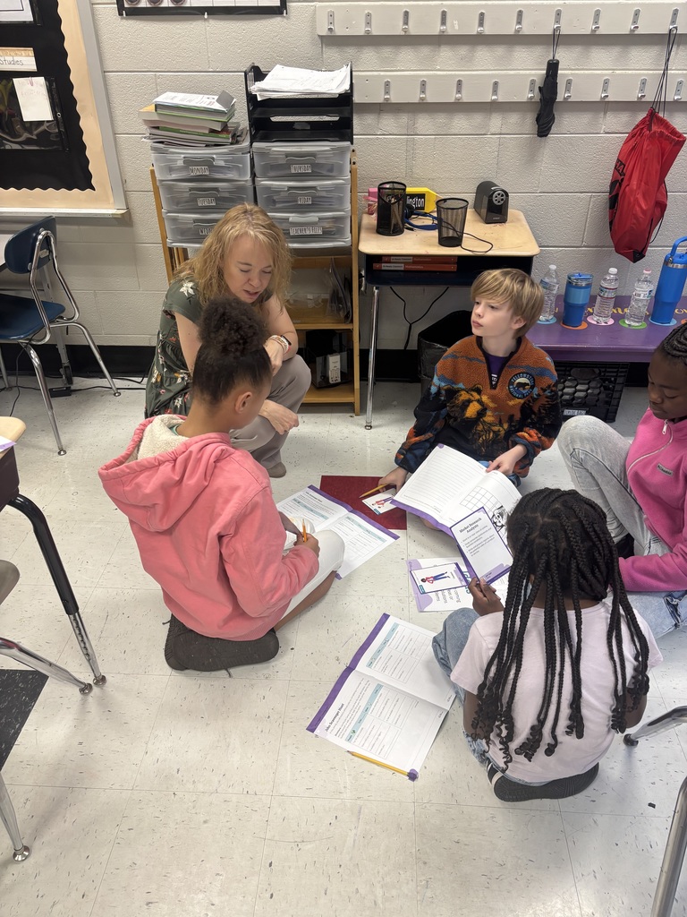 A teacher with blonde hair crouches on the floor interacting with four students seated on light-colored tile in a classroom learning activity. Two girls with dark hair wear pink hoodies, one with braids wearing a light pink shirt. A boy with blonde hair wears a patterned fleece jacket, and another girl in a pink hoodie is partially visible on the right. Students have open textbooks and worksheets with pencils visible. The student with braids works on "Week 1: Research Ready?" materials covering asking questions, finding information, using evidence, and sharing what you learn. A "Skill-Builder Chart" is visible on the floor