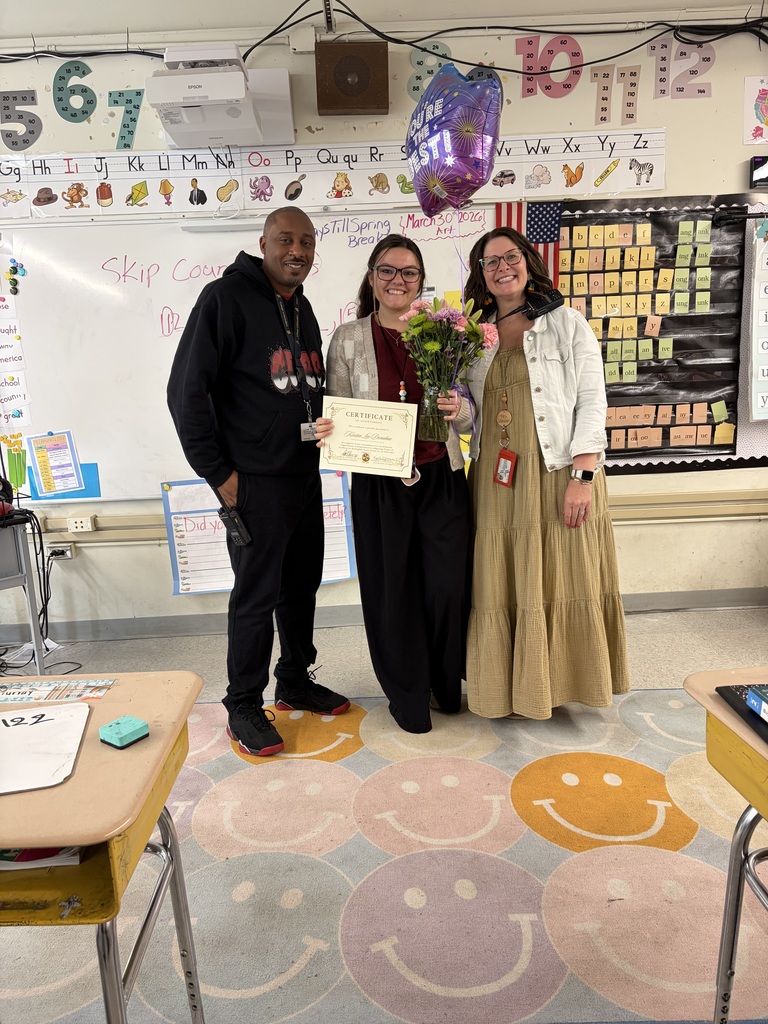 Three people smile together in a brightly lit classroom during a celebration. A man stands on the left, a woman in the center holds a certificate and a bouquet of flowers, and another woman stands on the right. The woman's certificate reads "CERTIFICATE This Certifies That Kristen Joy Gardner has been presented for outstanding achievement in Excellence in [Teaching/Learning] Presented this 24th day of March, 2026" with signature lines. A purple balloon with a star/fireworks design reading "YOU'RE THE BEST!" is visible nearby.