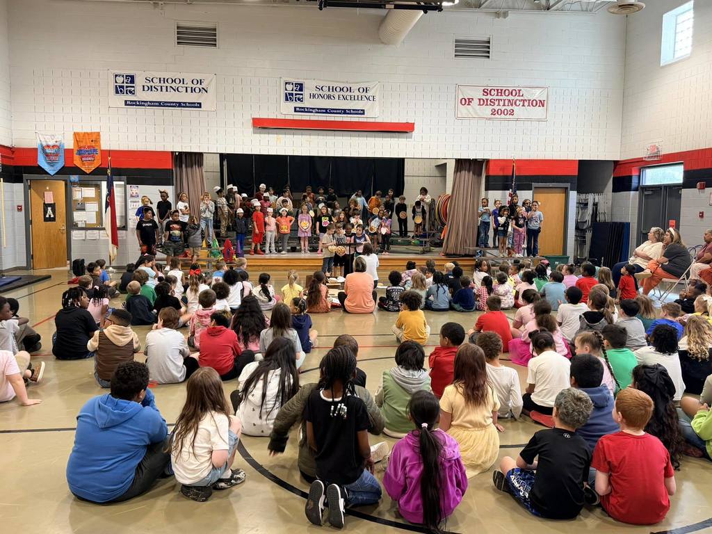 A school event takes place in a gymnasium where a large group of elementary school children are seated on the floor facing a raised stage. Several adults are seated in chairs on the right side, observing the event. The gymnasium features high ceilings, red and black painted walls, and a wooden floor with court lines. Banners on the wall above the stage recognize "School of Distinction" and "School of Honors Excellence" from "Rockingham County Schools." A North Carolina state flag is visible to the left of the stage.