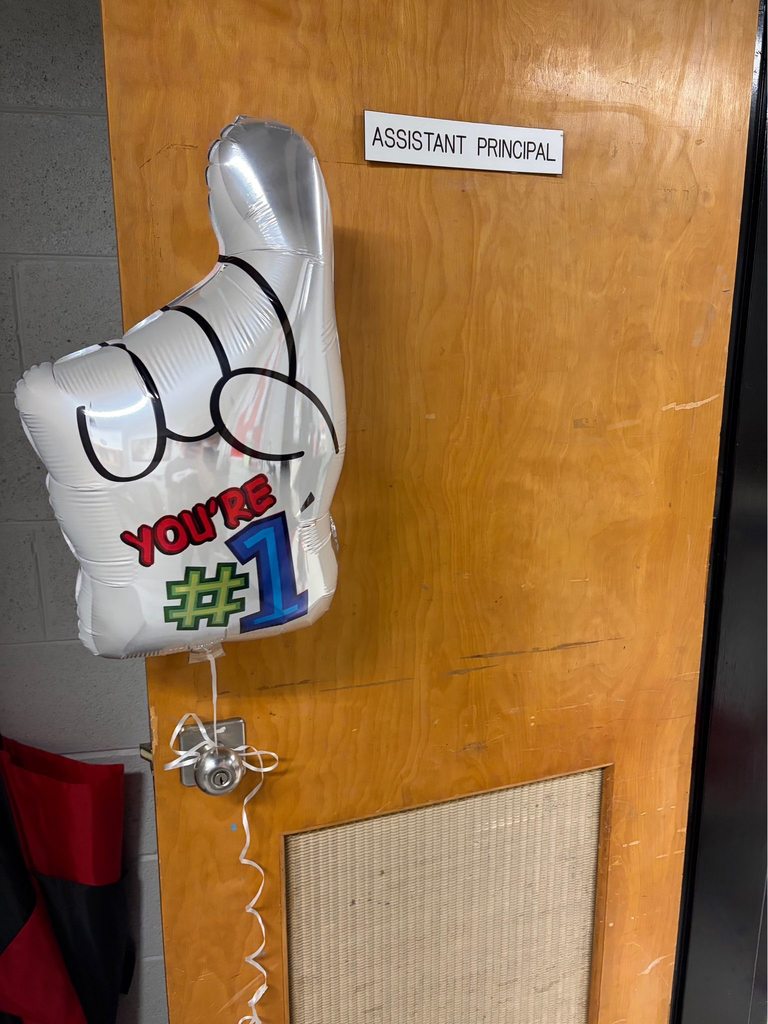 A light brown wooden door with a round silver doorknob is decorated with a silver Mylar foam finger balloon tied to the knob with white curly ribbon. The balloon displays black lines indicating fingers and text reading "YOU'RE #1" in red, green, and blue. A white rectangular sign is affixed to the door above the doorknob reading "ASSISTANT PRINCIPAL YOU'RE #1." A grey concrete block wall is visible to the left of the door, with a red and black object partially visible on the floor. The bottom right of the door shows a textured panel.