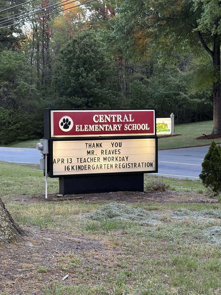 A roadside sign for "Central Elementary School" displays school announcements and recognition. The top part of the sign features red lettering with white text and a paw print logo next to the school name. The lower part is a lighted message board displaying announcements reading "THANK YOU MR. REAVES APR 13 TEACHER WORKDAY 16 KINDERGARTEN REGISTRATION." Numerous trees are visible in the background, with a road to the left of the sign. A small commercial sign in green and yellow is partially visible to the right. Grass and fallen or cut leaves appear in the foreground.