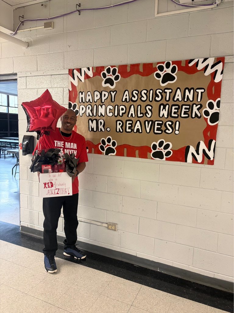 A man stands in a school hallway smiling while holding gifts to celebrate Assistant Principals Week. He wears a red t-shirt printed with "THE MAN THE MYTH," black pants, and dark-colored sneakers. In his left hand, he holds a red star-shaped balloon. In his right hand, he holds a white gift box filled with red-themed items including a red Sharpie marker and other gifts, decorated with black tissue paper. A white sign on the box reads "TO an AP WHO is RED iculously AMAZING!" Behind him, a large bulletin board banner decorated with black paw prints and red wavy borders on a brown background displays "HAPPY ASSISTANT PRINCIPALS WEEK MR. REAVES!" The hallway features white brick-patterned walls, a black baseboard, and light-colored tiled flooring. In the background, an open doorway reveals a cafeteria or common area with tables and chairs.