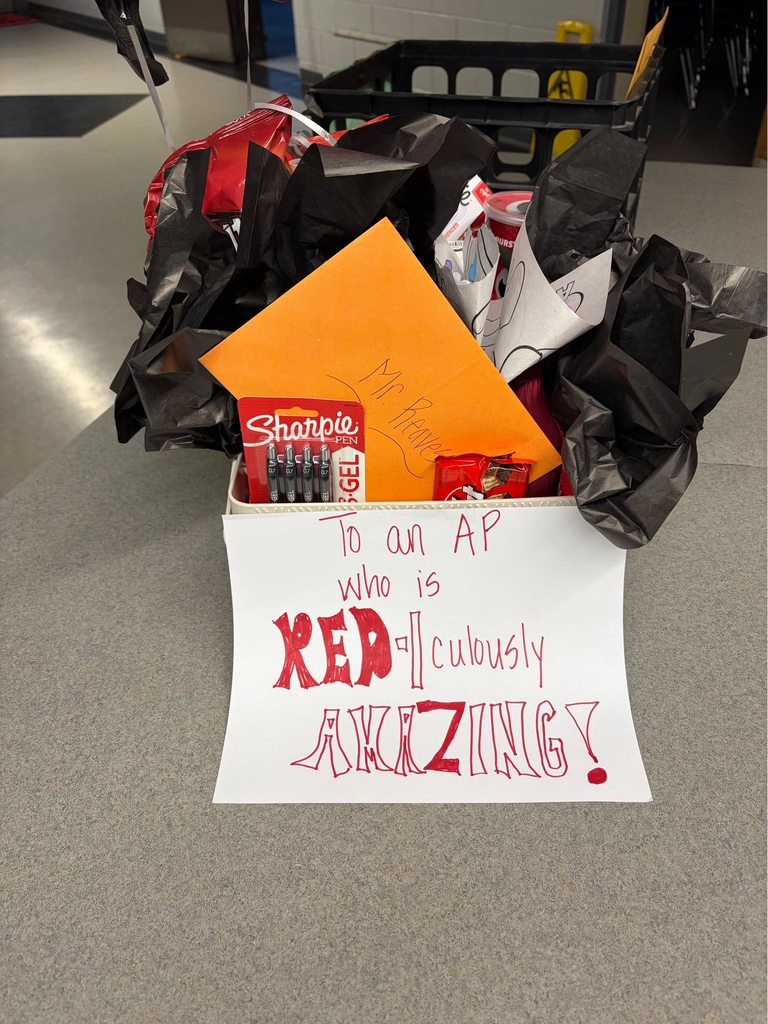 A gift box is filled with various red-themed items and office supplies, decorated with black tissue paper. An orange envelope addressed "Mr. Reaves" is prominently displayed within the box. A package of Sharpie 0.7 GEL PENS, a Kit Kat chocolate bar, and other red-packaged snacks are visible. A white sign attached to the front of the box displays the message "To an AP who is RED-iculously AMAZING!" The box rests on a speckled gray countertop with crates or shelves visible in the blurry background.