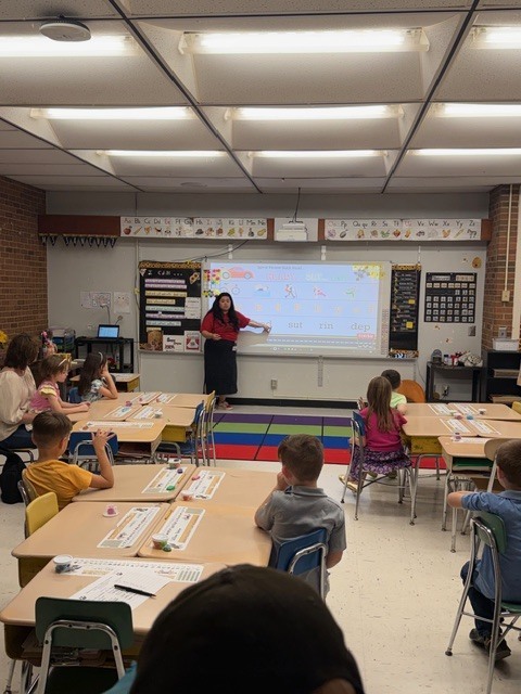 A classroom scene captures a female teacher standing at the front pointing to an interactive whiteboard displaying educational content. Students are seated at individual desks arranged in rows facing the front, with worksheets and papers visible on their desks. An additional adult is seated with students on the left side of the room. The classroom features fluorescent lighting and a tiled ceiling. A brick wall is visible on the left. Educational posters and charts adorn the walls surrounding the whiteboard. A colorful striped rug in red, blue, green, and purple is laid on the floor in front of the whiteboard. A ceiling-mounted projector casts the image onto the whiteboard.