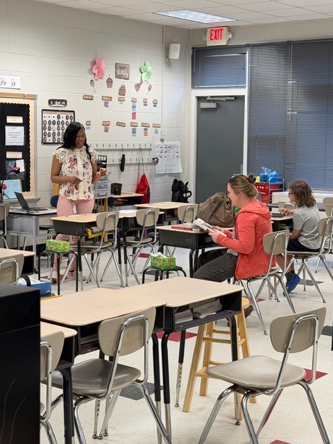 A brightly lit classroom scene shows an African American woman, likely a teacher, standing in the center-left foreground smiling and gesturing as if speaking. She wears a floral short-sleeved blouse and light pink pants. A silver laptop rests on a desk to her left. The classroom features light-colored cinder block walls with educational displays and with decorative paper cutouts in pink and green and brown acorn-shaped cutouts with colorful labels. Hooks on the wall hold various items including a red bag. Several student desks with attached chairs are visible in rows. A woman with light brown hair in a red/orange long-sleeved top sits at a desk in the mid-ground reading a book. Another individual in a grey t-shirt sits at a nearby desk. A light wooden stool and tissue boxes are positioned near desks. The floor features light-colored tiles with red and black accents. On the right side, an emergency EXIT sign is mounted above a grey door with horizontal blinds visible above. Ceiling tiles and fluorescent lighting fixtures are visible throughout. A white speaker is mounted on the wall above the door. Desk labels showing "No 17" and "No 20" are visible on the front edges of desks.
